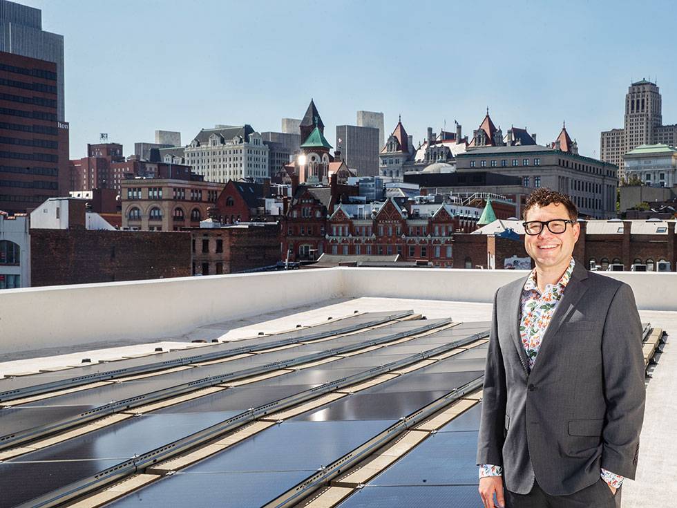 Brendan Woodruff stands on a rooftop next to solar panels with a view of the city in the background.
