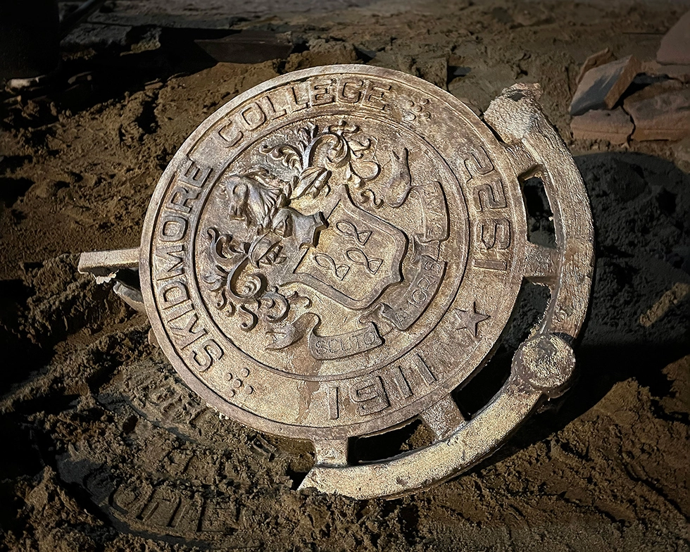 A newly cast Skidmore College bronze seal, detailed with the college crest and motto, rests in a bed of sand inside a metal forge workshop.