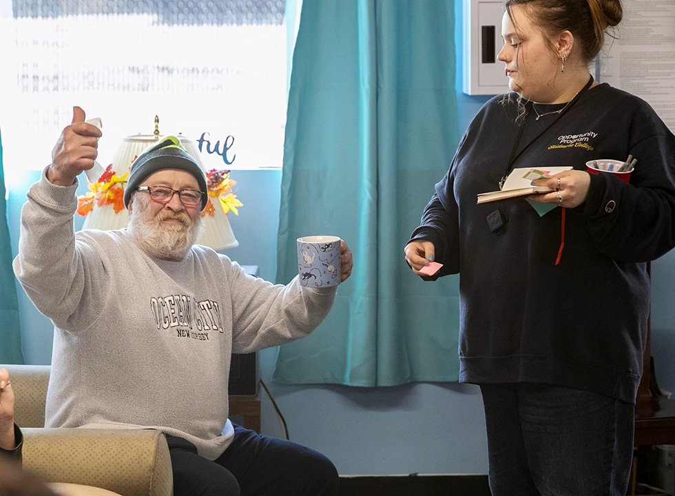 Skidmore student engaged in community service, speaking with a local resident holding a coffee mug and giving a thumbs up at a community organization.