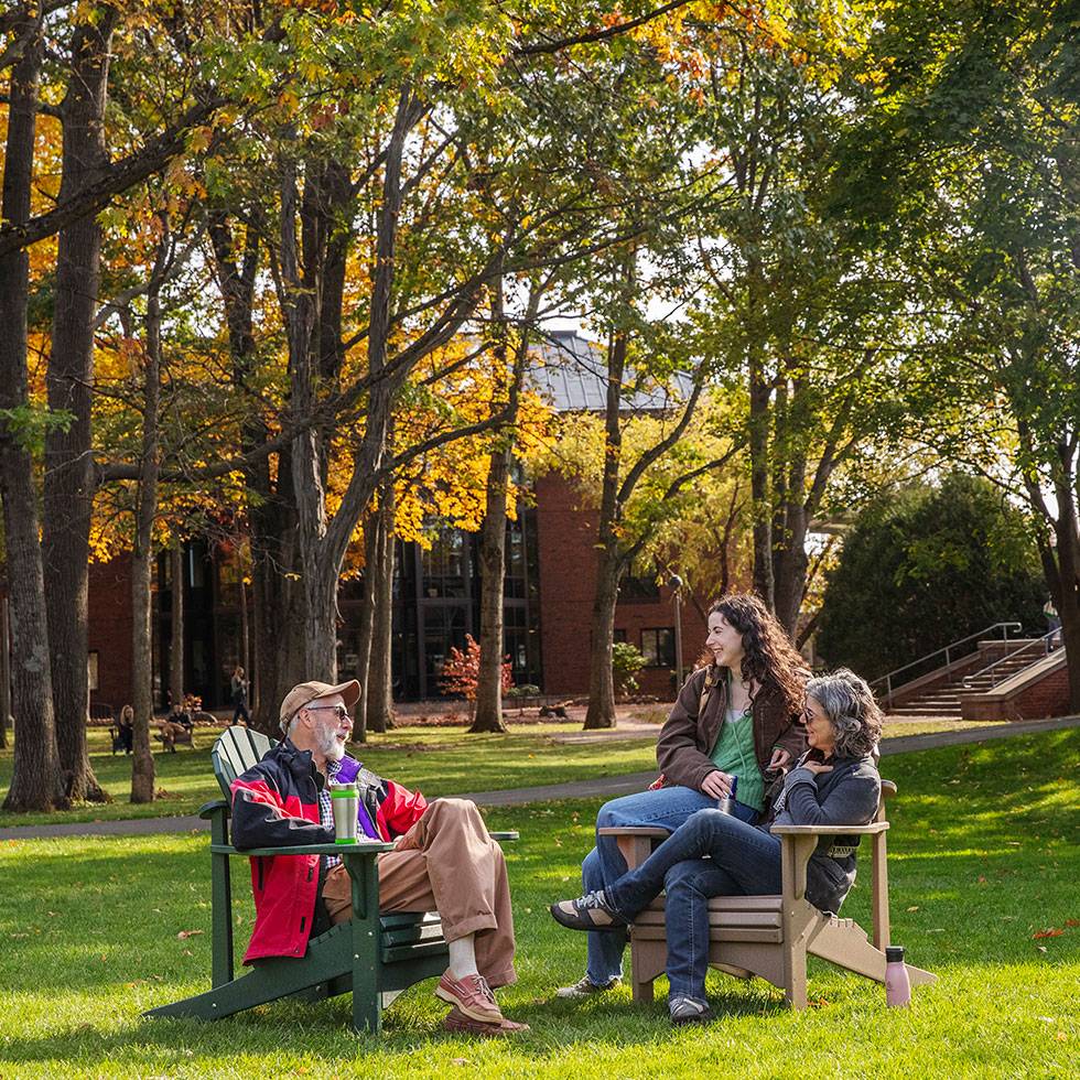 A family sits in adirondack chairs with beautiful fall foliage and the library in the background