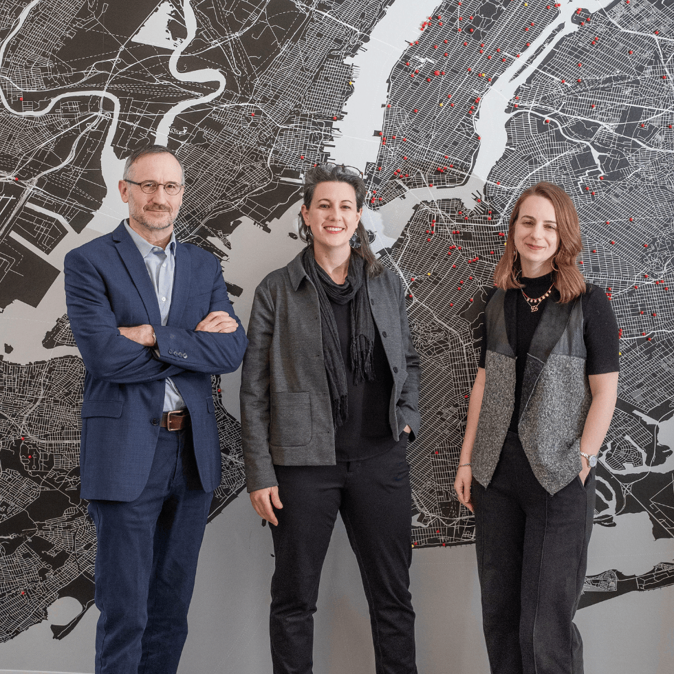 Three alumni posing in front of a map of NYC with pins on architecture projects their firm has worked on.