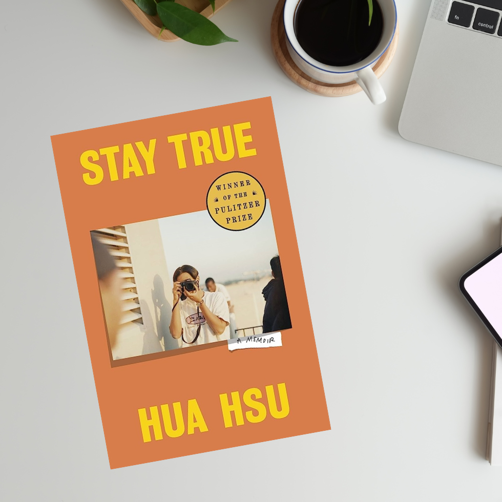A book, "Stay True" by Hua Hsu, lays flat on a desk with coffee and a laptop