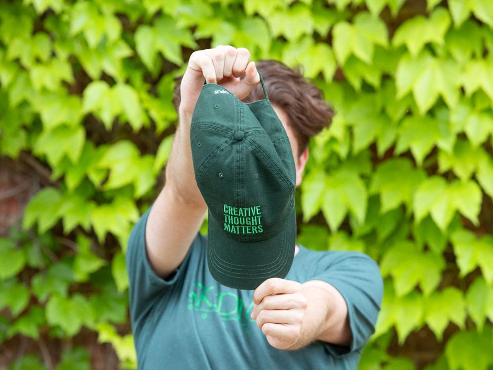 A person holds a Creative Thought Matters hat in front of their face with a a wall of green ivy in the background