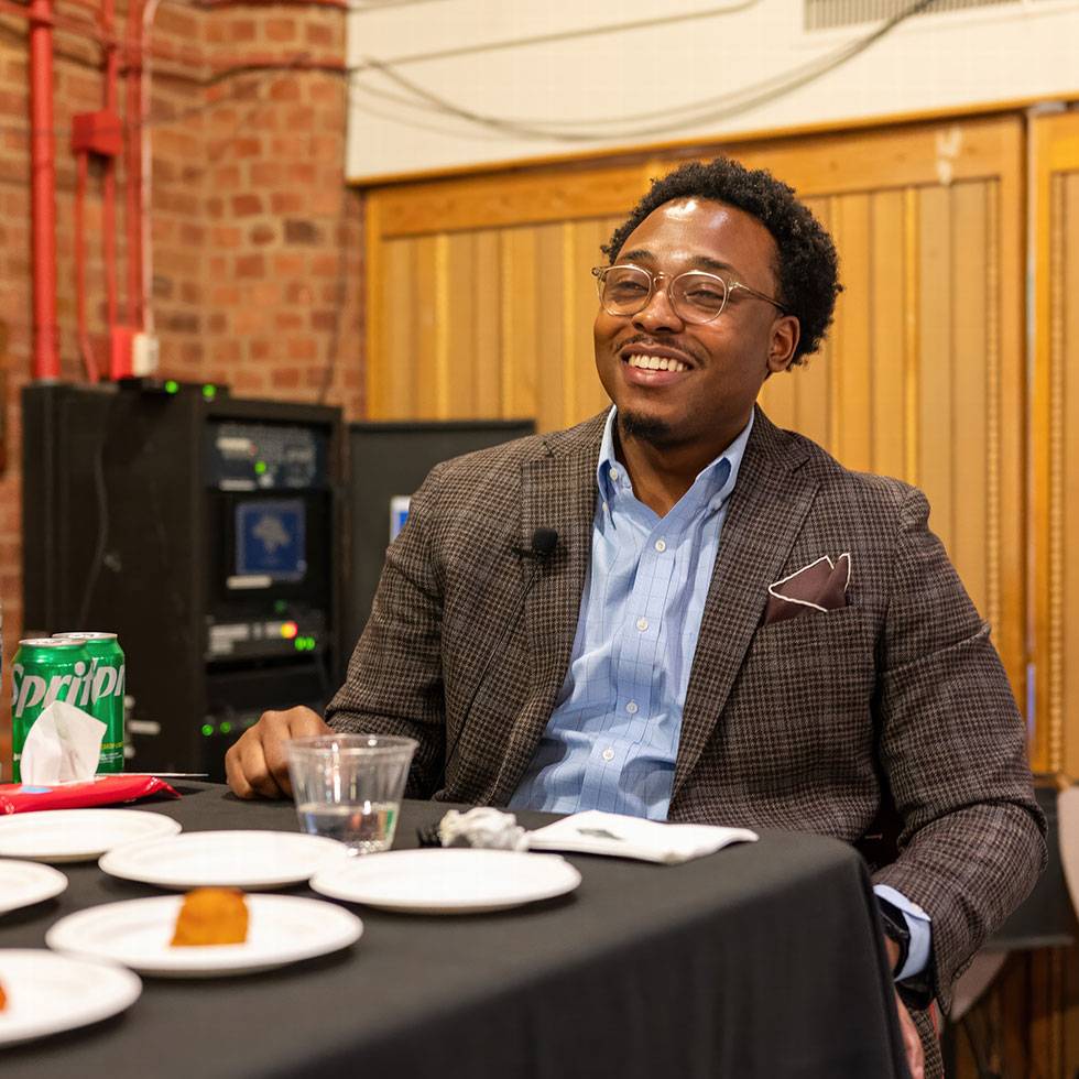 Nigel Smith sits smiling at a table wearing a plaid blazer and blue button-down shirt with white plates in front of him.