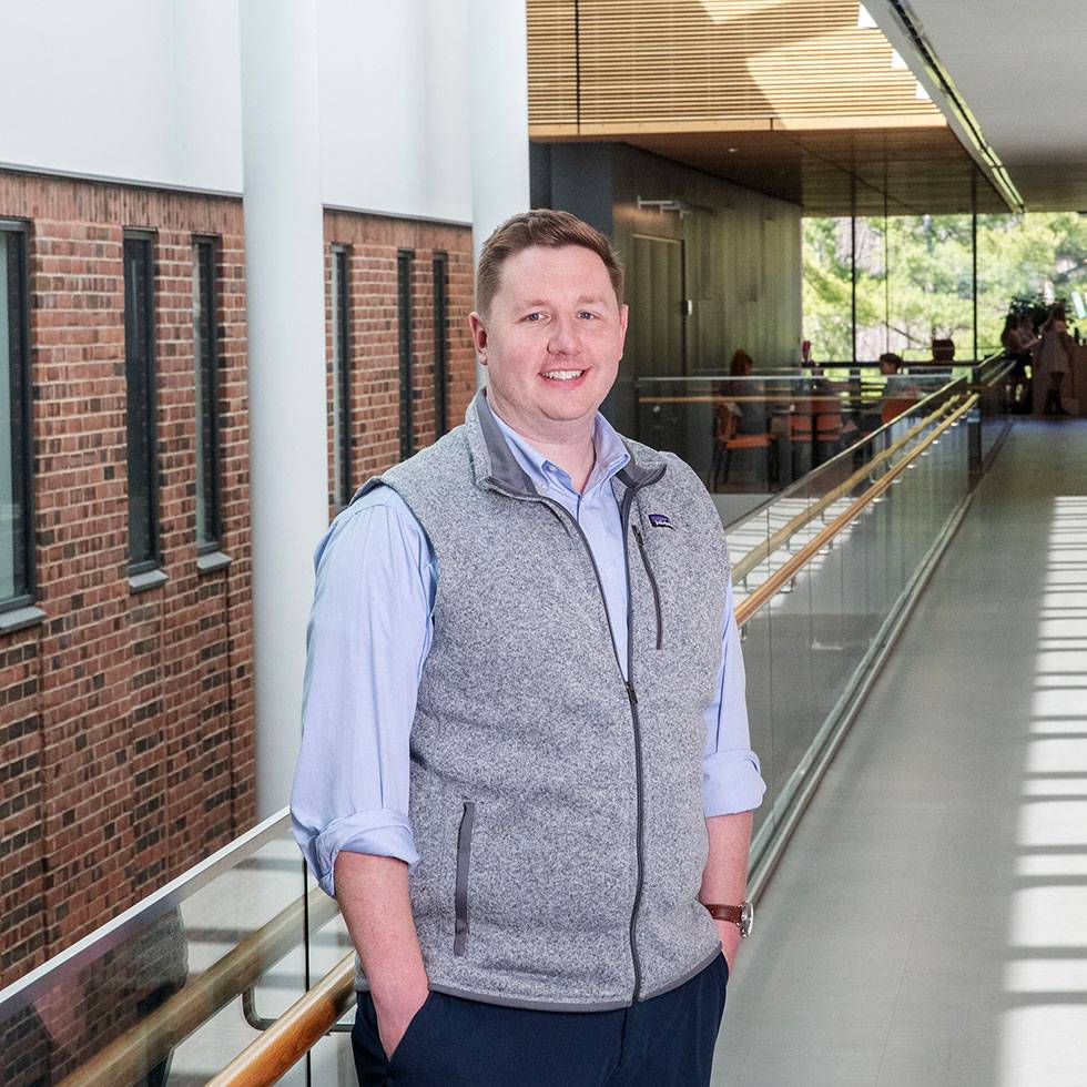 Matt Walsh stands on an interior balcony of the Billie Tisch Center for Integrated Sciences wearing a grey vest and blue button-down shirt, with his hands in his pockets. The background includes an interior brick wall with windows.