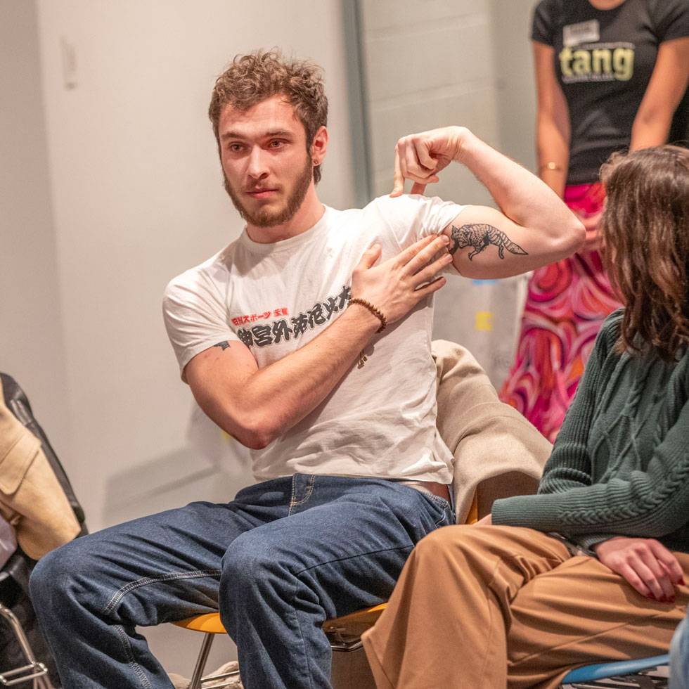 A student sits in a chair wearing a white T-shirt and jeans, holding their left arm up to show off a tattoo on their bicep that resembles a coyote.