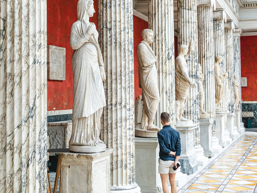 A person stands in the interior hall of the Ny Carlsberg Glyptotek in Denmark with Roman and greek marble sculptures statues and columns. 
