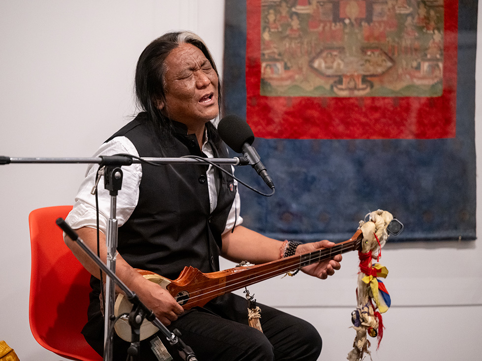 A guest musician performs on a traditional stringed instrument while singing into a microphone during a campus cultural program, with artwork displayed behind the performer.