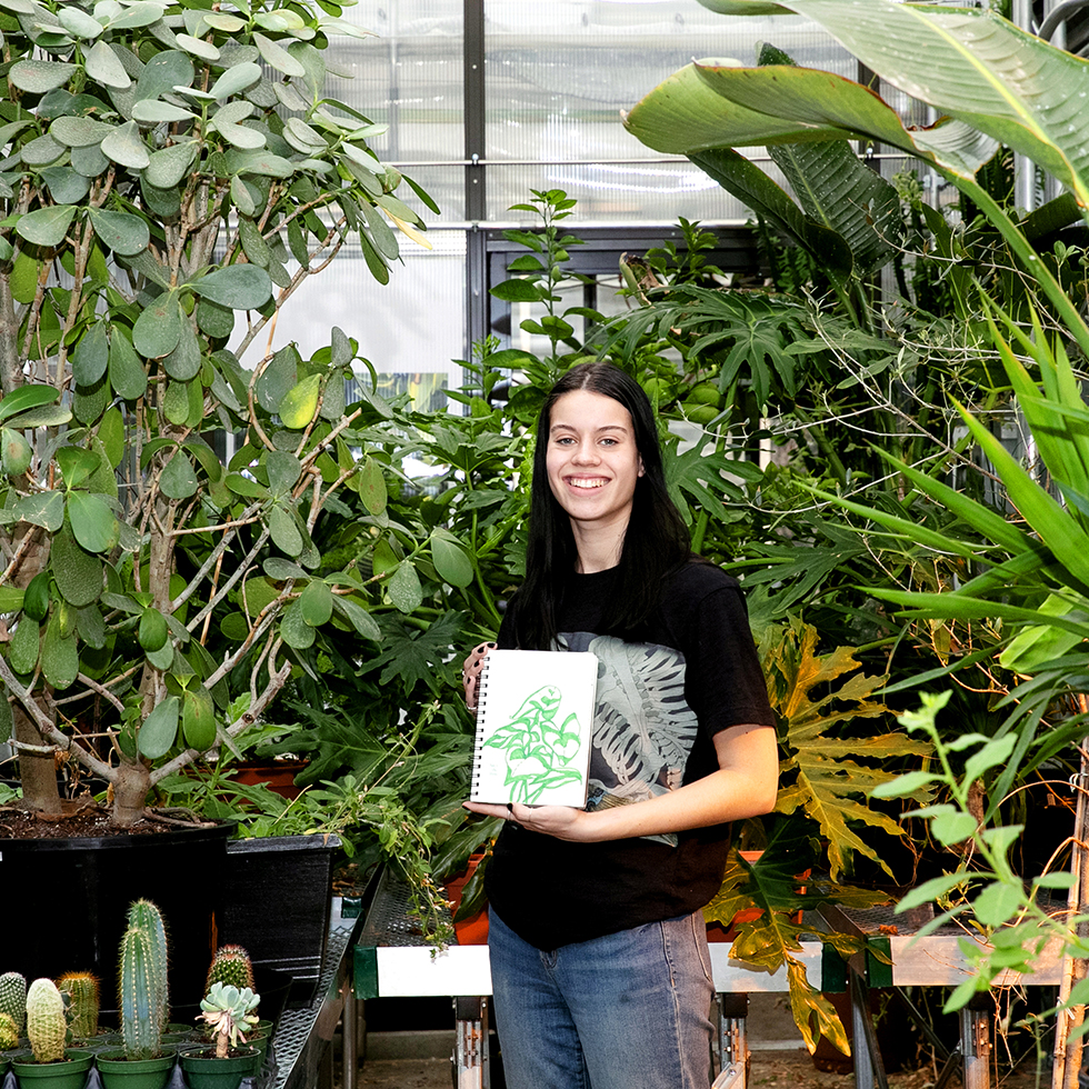 Person holding a sketchbook with a plant drawing, standing among various green plants in a greenhouse.