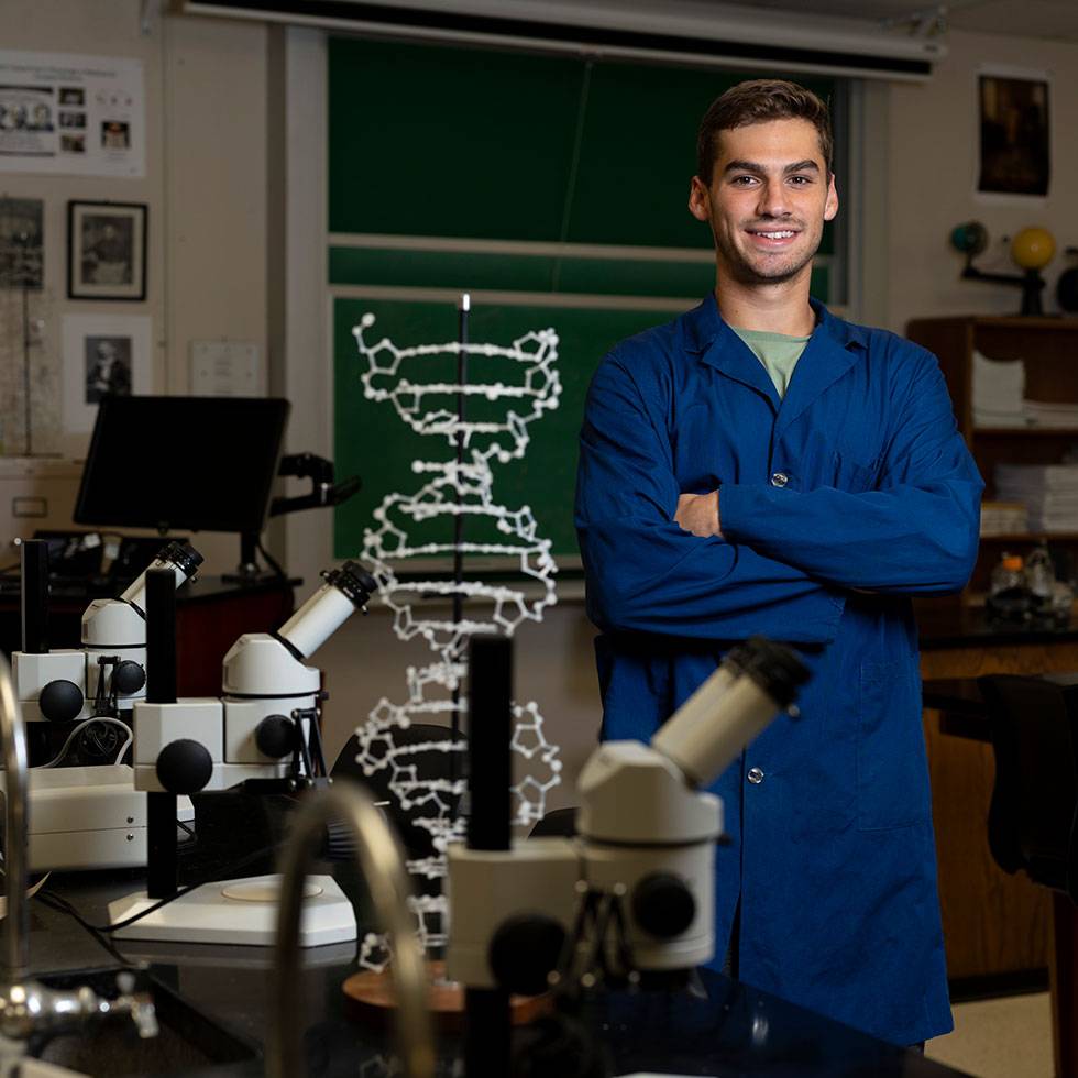 Student stands with crossed arms in a blue lab coat surrounded by microscopes and other scientific equipment