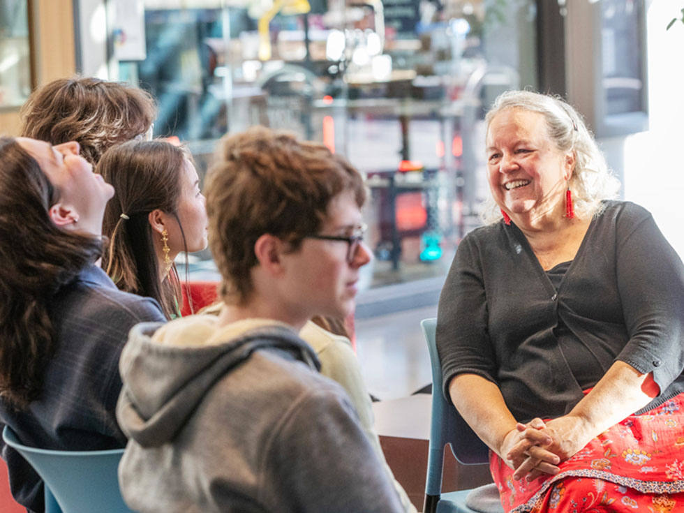 A smiling woman sits in conversation with a small group of students in a brightly lit indoor space. The group appears relaxed and engaged, with some students laughing and others listening attentively.