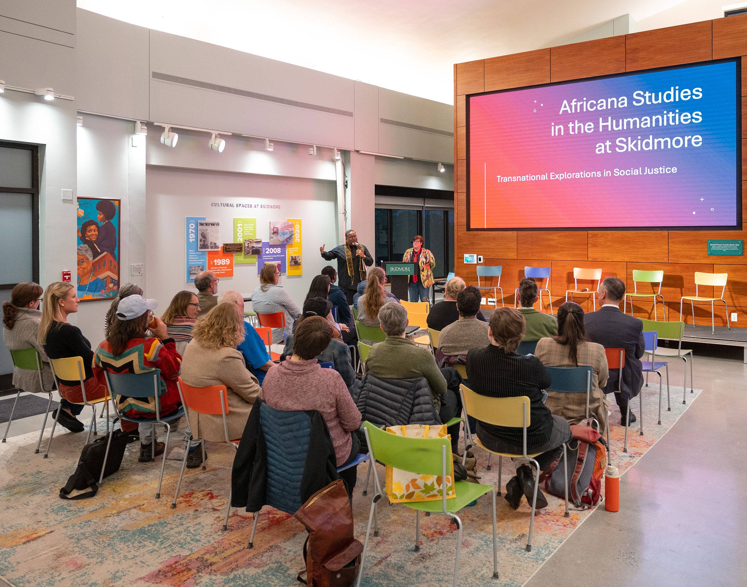 An audience sits in colorful chairs listening to two presenters at the front of the room. The background includes a powerpoint slide that reads "Africana Studies in the Humanities at Skidmore", and fades from orange to blue.