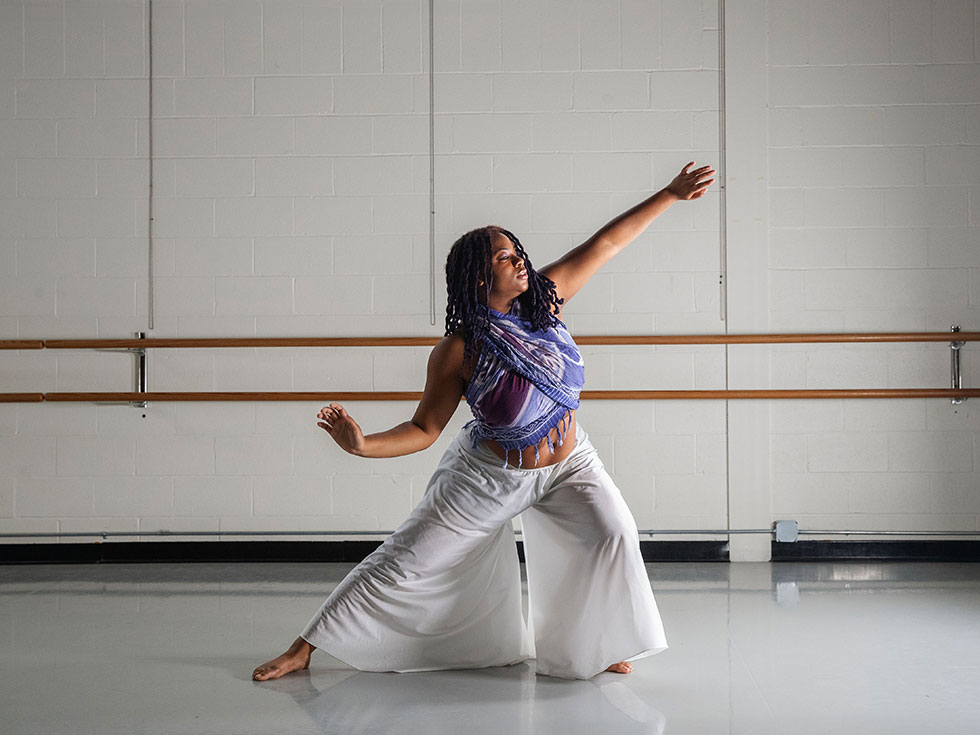 A dancer poses in a dance studio, one arm extended overhead and the other lowered, wearing a patterned blue-purple wrap top and light-colored pants, with ballet barres and a softly lit wall in the background.
