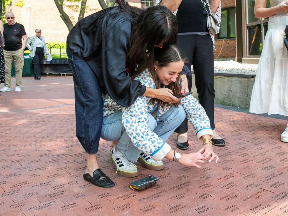 A family member curls around student crouched on the ground, both taking a photo with their phones of the students engraved brick