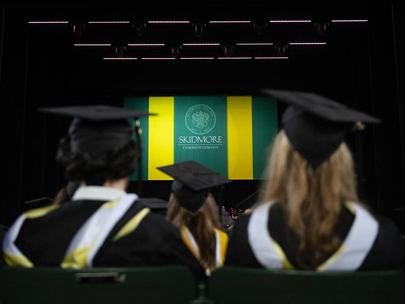 Dark ampitheater with a screen that alternates yellow and green and reads "Skidmore Commencement" frame in between two students backs wearing graduation regalia