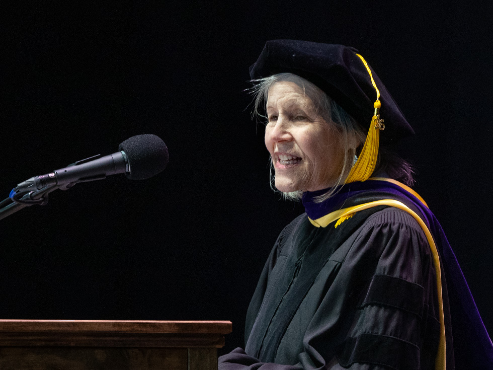 JoAnne Manson wears black academic regalia and a doctoral cap while addressing the Skidmore audience from the podium.