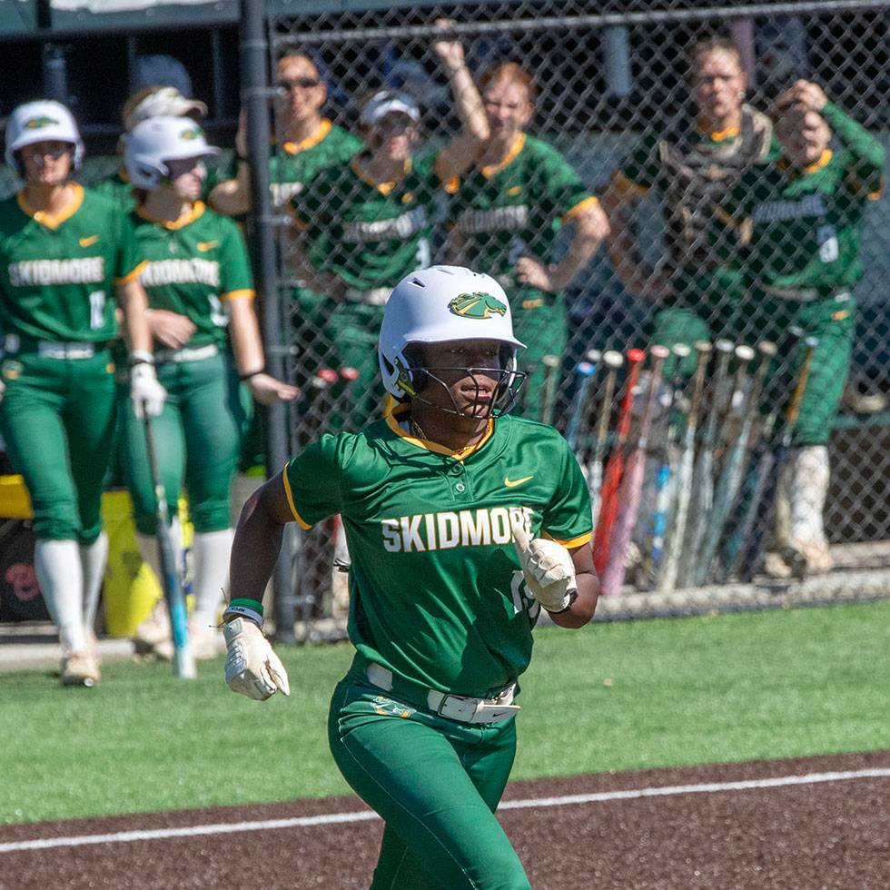 Skidmore softball player running to first base in their uniform with teammates cheering in the dug out.