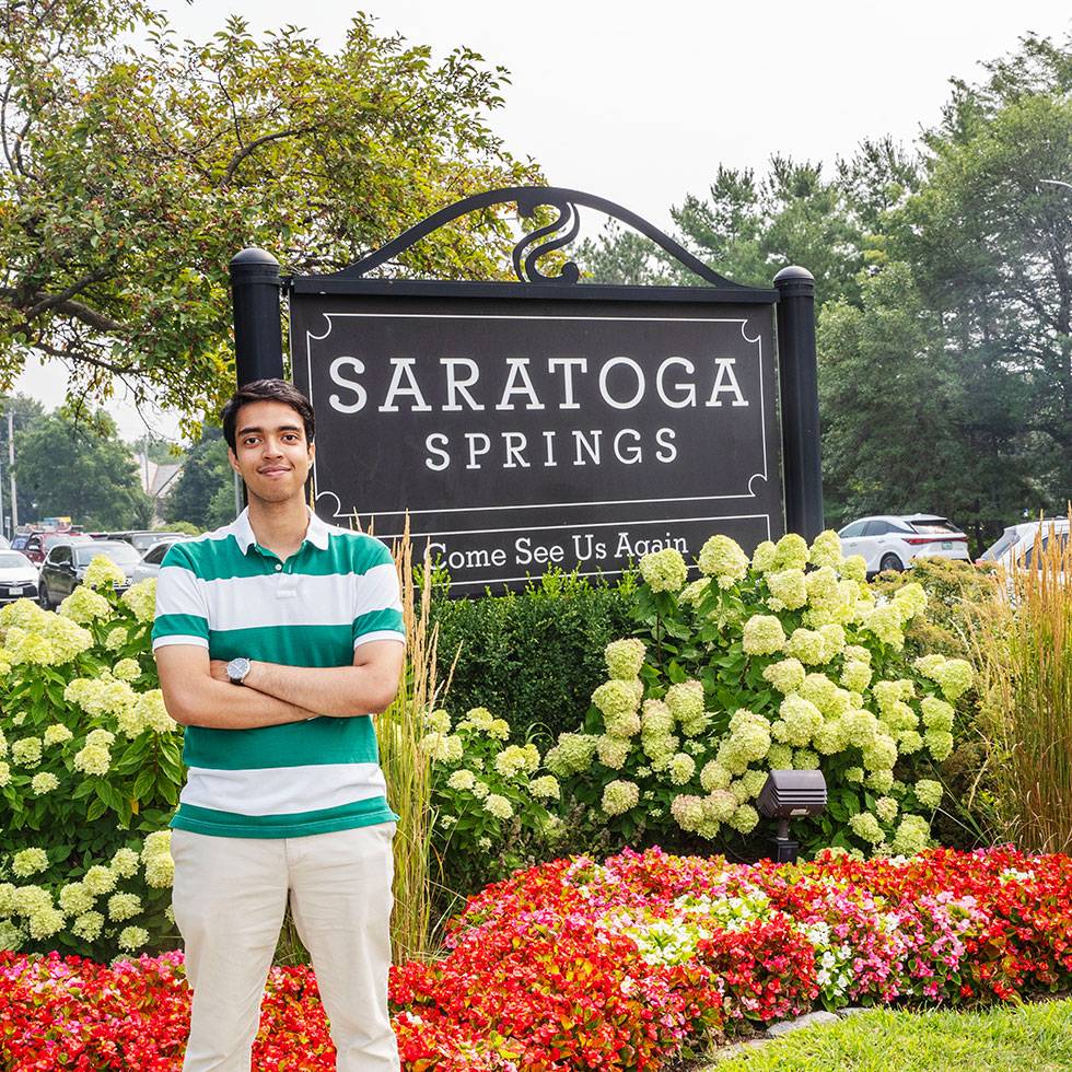 A young man stands in front of a Saratoga Springs sign wearing a green-and-white striped polo shirt and light-colored pants. He faces the camera with his arms crossed. 