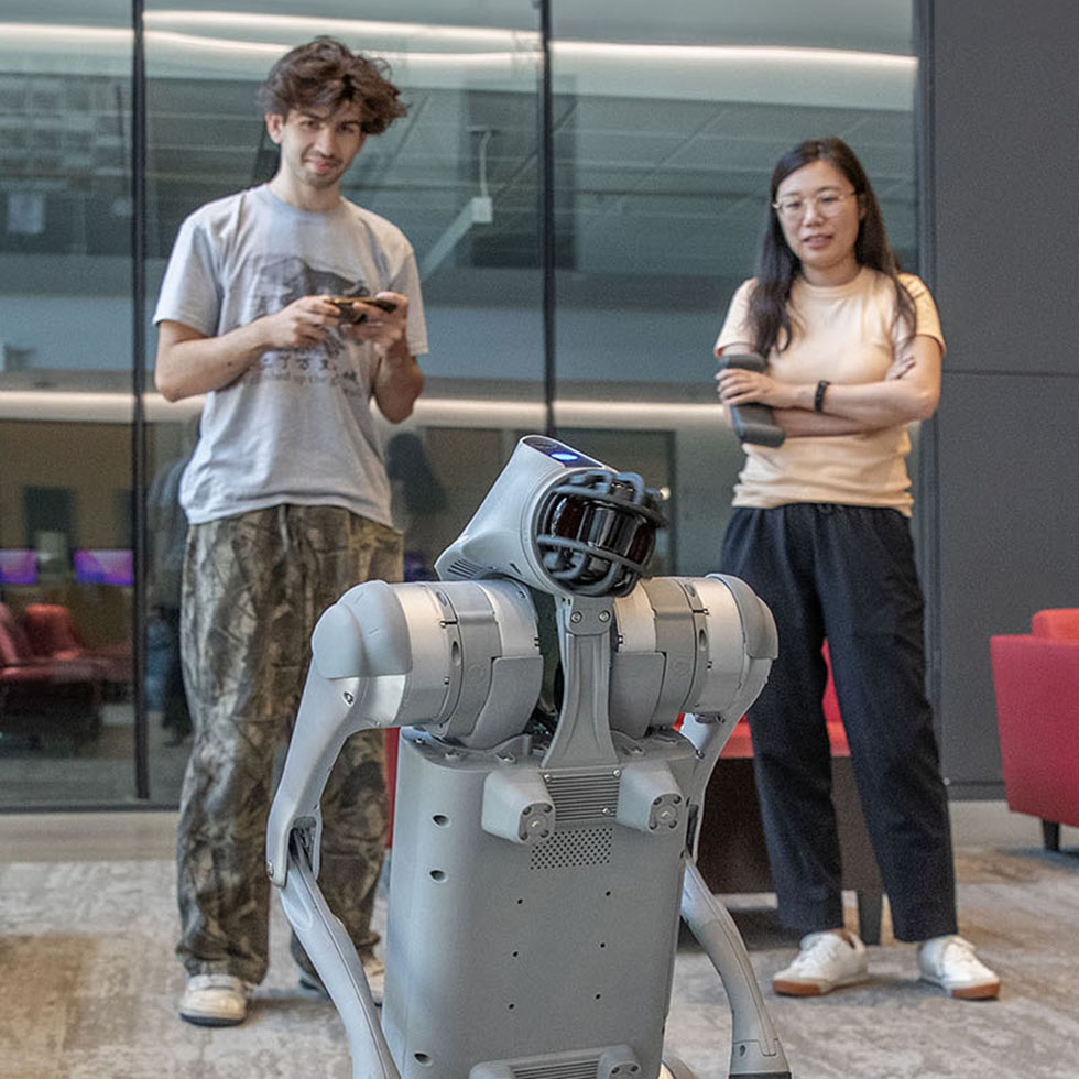 Two researchers observe and operate a humanoid robot inside a modern campus building. One holds a controller while the other watches with arms crossed, as the robot stands in the foreground against a backdrop of glass walls and lounge seating.