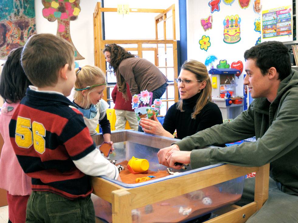 A professor and education student crouch down to a sensory play table to talk to three young children in Skidmore's Early Childhood Center