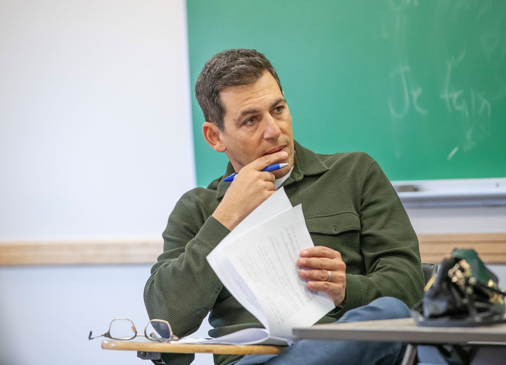 Stuart Zicherman sits at a desk with a green chalkboard in the background, holding a piece of paper in one hand and his other hand resting at his mouth holding a pen