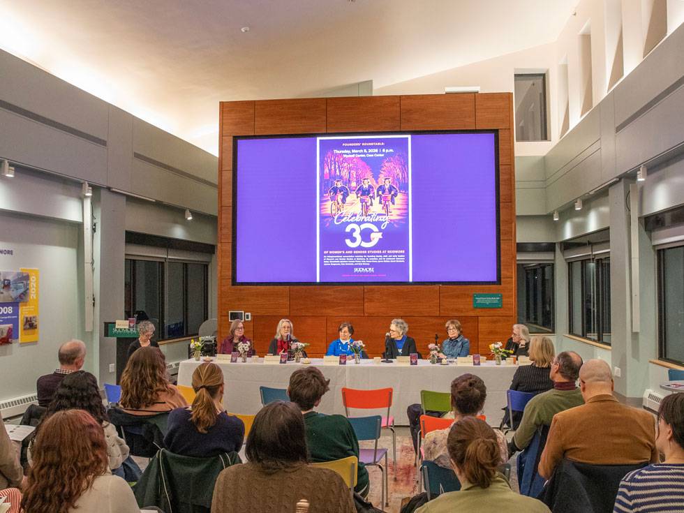 An audience sits in colorful seats, looking at a panel of speakers seated below a screen that shows a historic photo of three Skidmore students riding bikes and copy that says "Celebrating 30" for Skidmore's Gender Studies program.