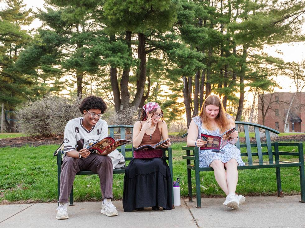 Three students sit on a green metal bench smiling, each holding a magazine. It is a sunny day and there are full green trees and grass in the background.