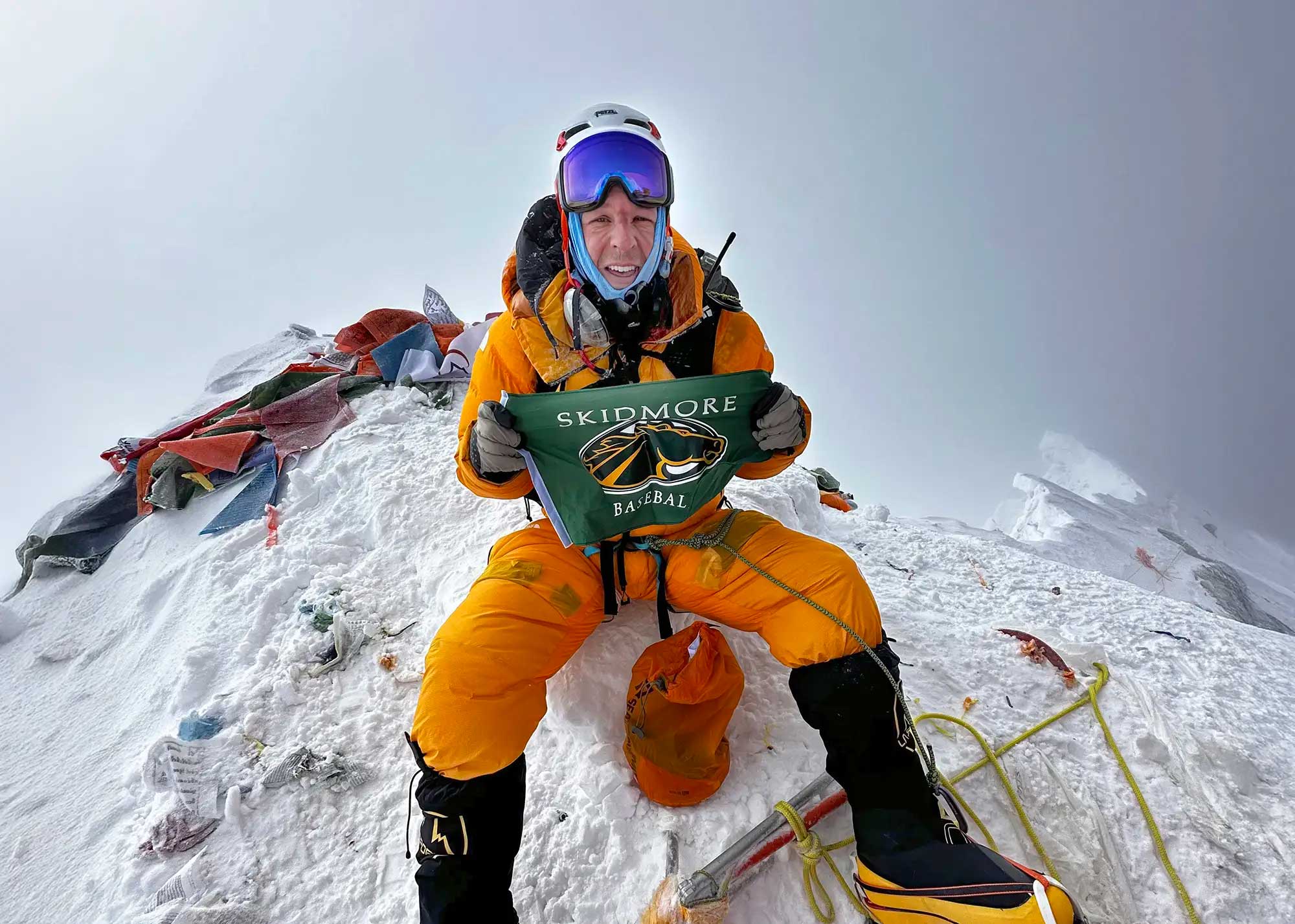 Mountaineer in bright orange cold-weather gear sitting on a snowy summit and holding a Skidmore Baseball banner, with climbing ropes and flags visible.