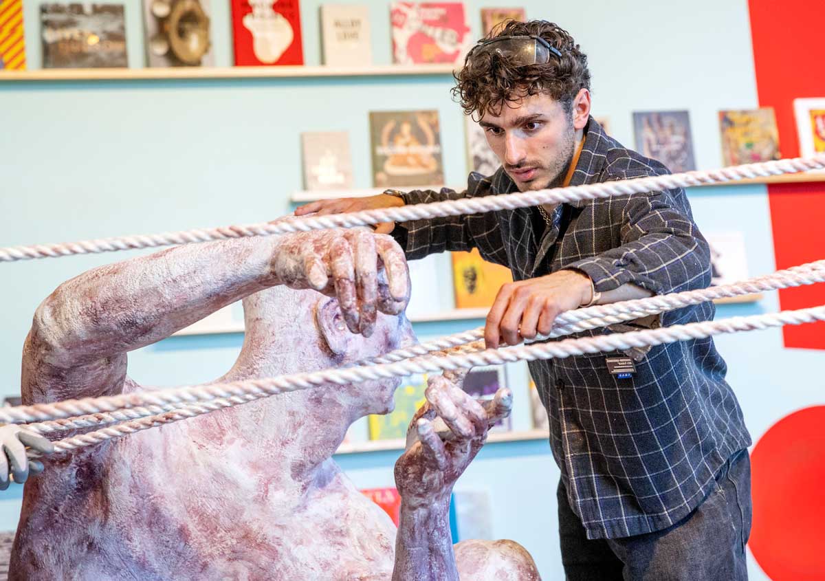 A student artist works on a large figurative sculpture surrounded by ropes in a gallery space.