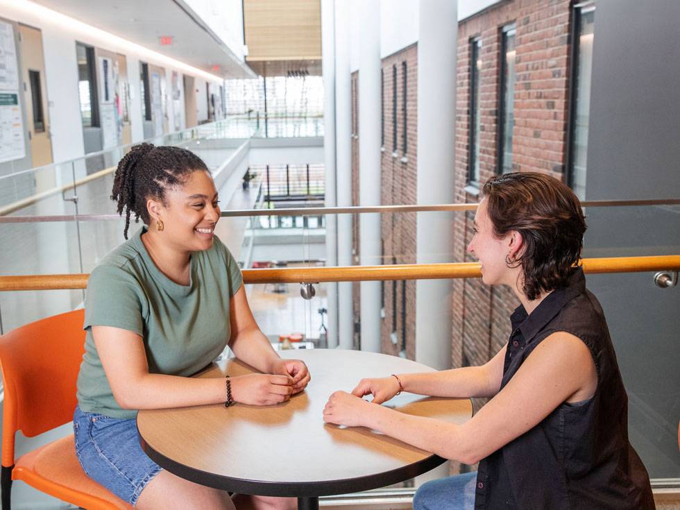 Two people sit on the upper interior balcony level of an academic building, smiling and having a conversation.