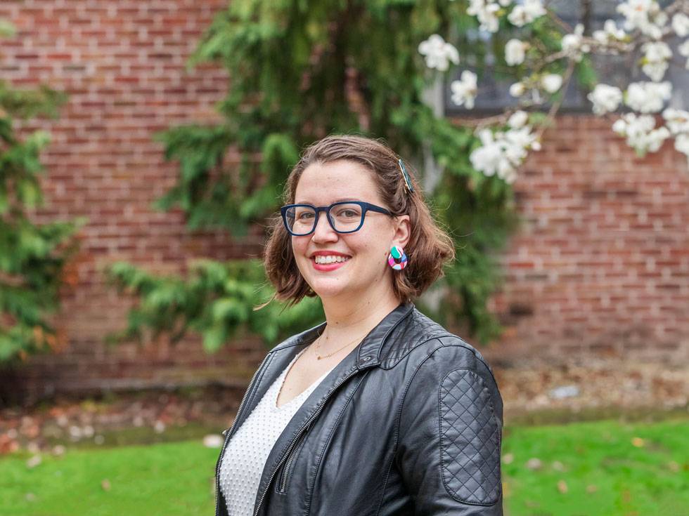 Sarah Faude '09 stands outside with glasses, a black leather jacket, and colorful earrings with a brick building in the background and green grass.