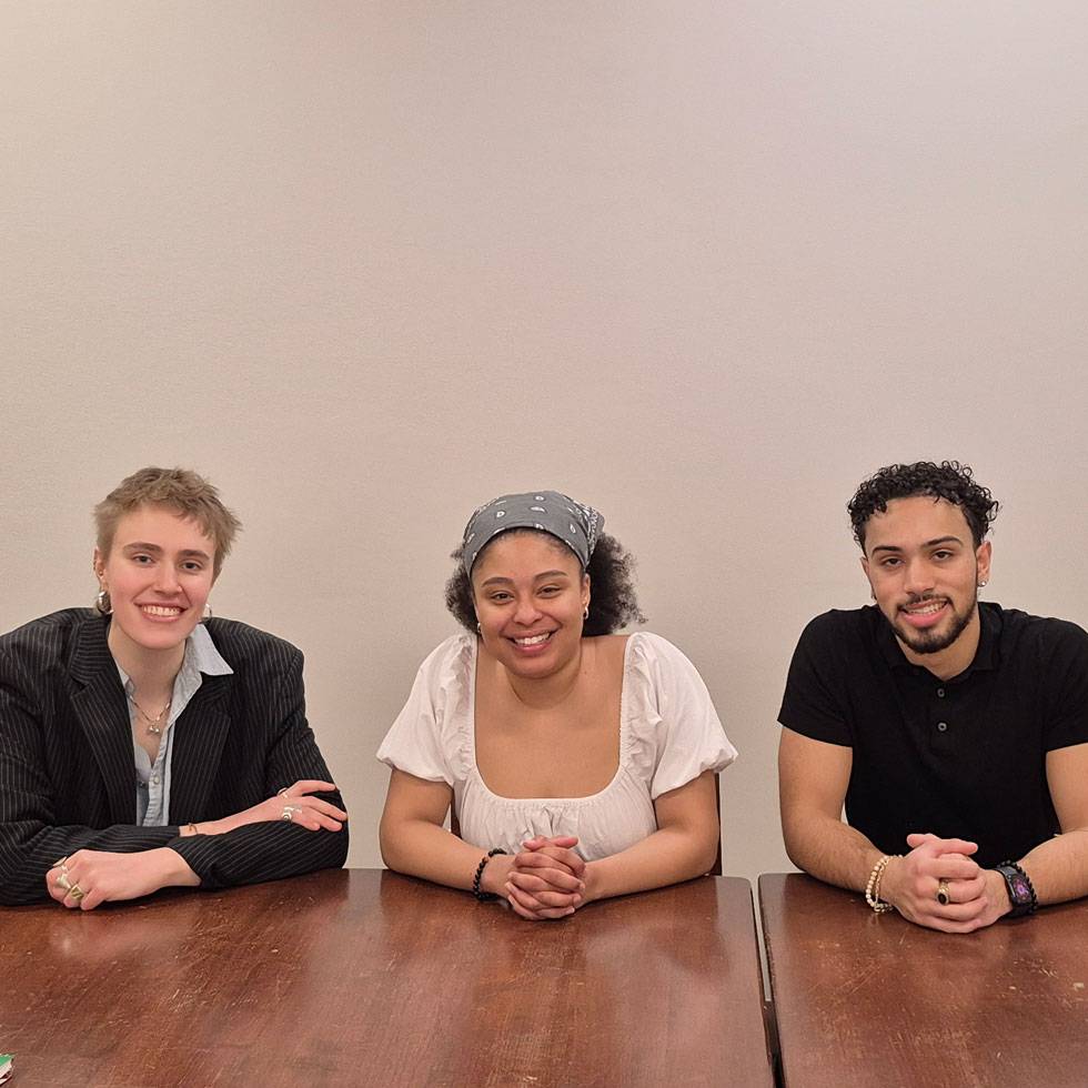 Three students sit at a table smiling, each with their hands clasped together in front of them resting on the table
