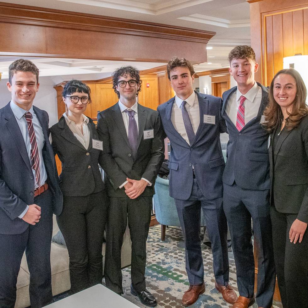 A group of students stand together in business attire, some with their arms around each other, smiling while they pose for their photo.