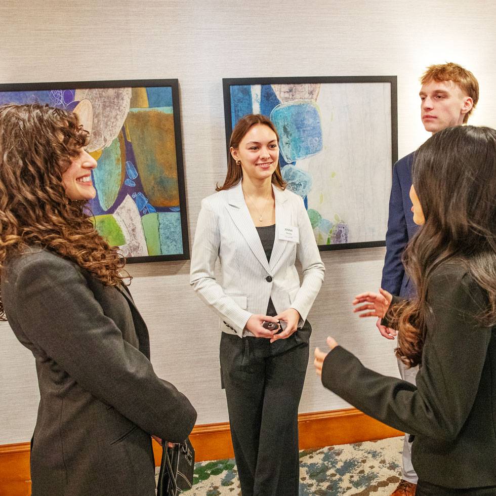 Four students stand in discussion wearing business attire, preparing to present at MB107