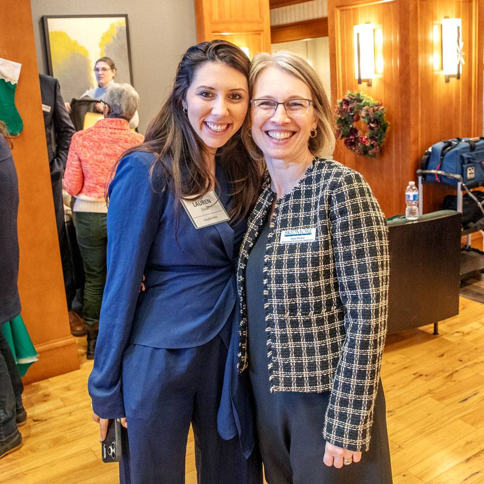A student a staff member stand with their arms around each other smiling. Both are wearing business attire and they are posing in a hotel lobby.