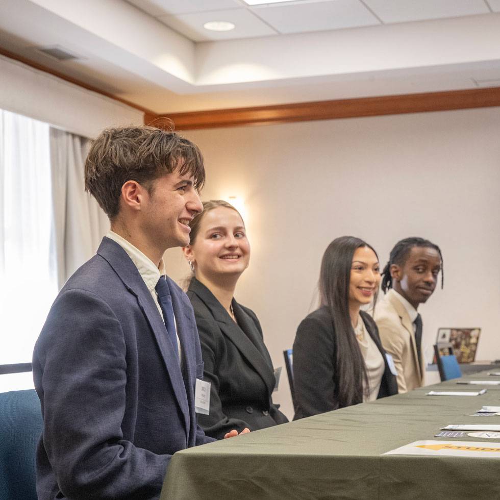 Four students sit at a conference table wearing business attire. The first student is smiling as they talk across the table to executives, who are not pictured.