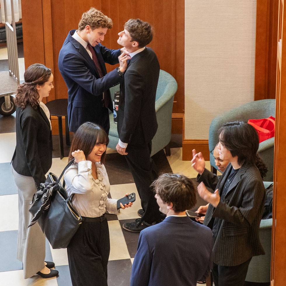 An over head view of students in business attire getting prepared to present at MB107. Some are talking to each other, while others are helping fix each others shirt collars.