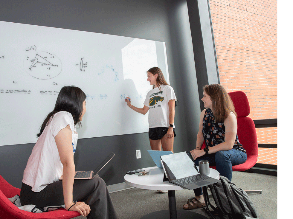 A student writes on a white board in a study space in the Billie Tisch Center for Integrated Science while another student a professor sit in red chairs watching