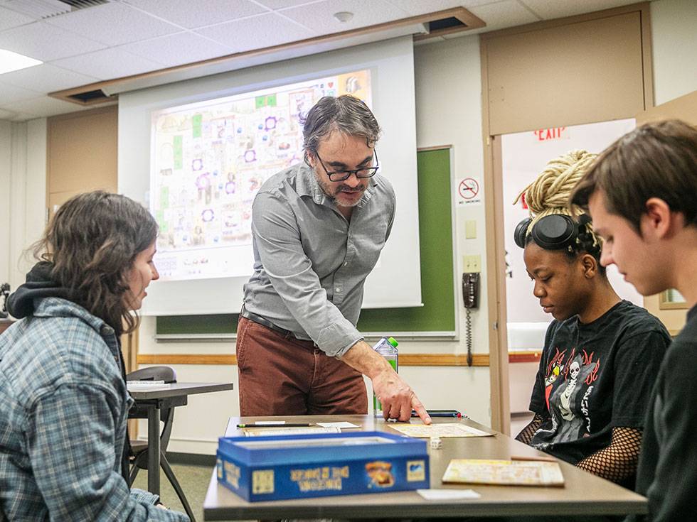 A professor guides a small group of college students through a tabletop game exercise in a classroom, using the game to illustrate key concepts projected on a screen behind them.