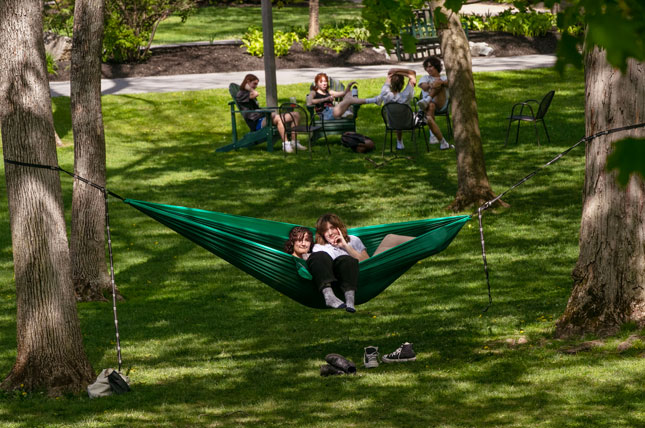 Two students relax together in a hammock strung between trees on Skidmore College’s campus while other students gather nearby on a fall day.