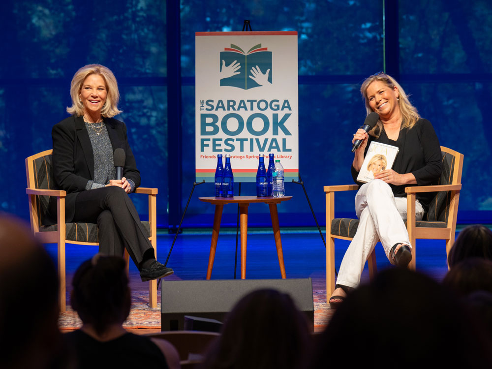 Joan Lunden and Julie Chapman sit on the stage of Zankel Music Center with a Saratoga Book Festival sign between them.