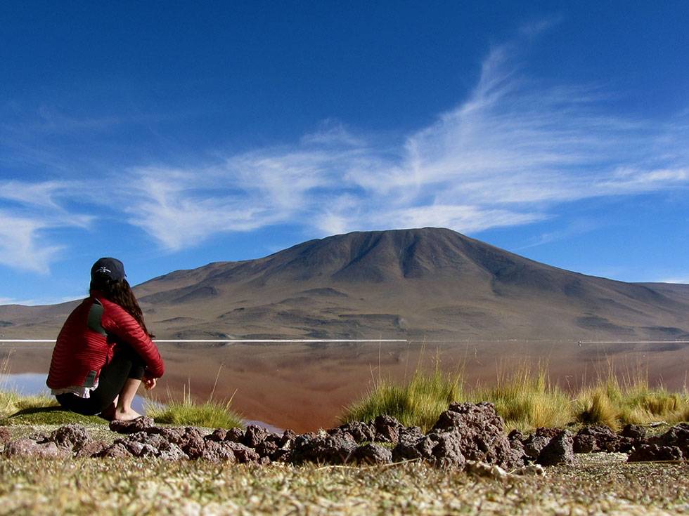 A student looks out over a lake with a mountain in the background on a blue sky day