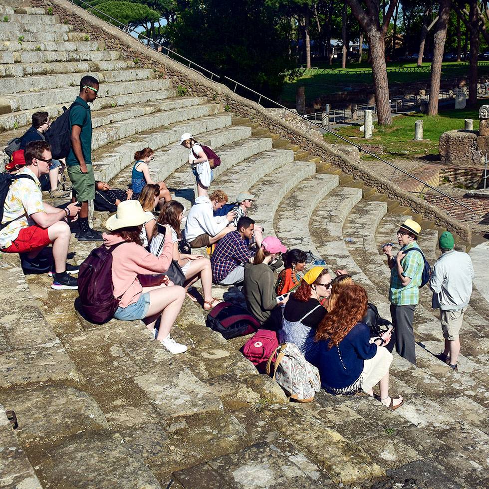 A group of students sit in a outdoor classical Roman amphitheater while listening to professors