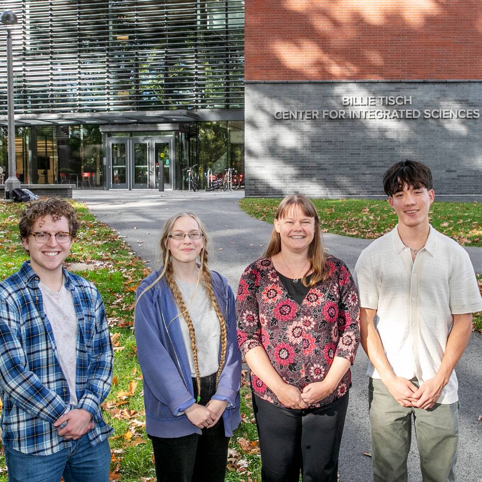 Professor Becca Johnson stands outside of Skidmore's Billie Tisch Center for Integrated Sciences posing with two students on her right and one on her left. It is a sunny day and you can see the science building in the background.clin