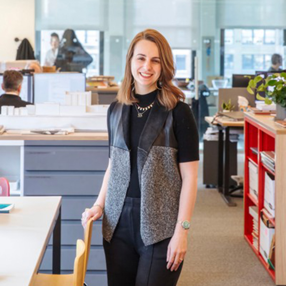 Sara Brandt stands in the architecture firm she works at. She is smiling, wearing a black and grey vest, and has her right hand resting on a wooden chair.
