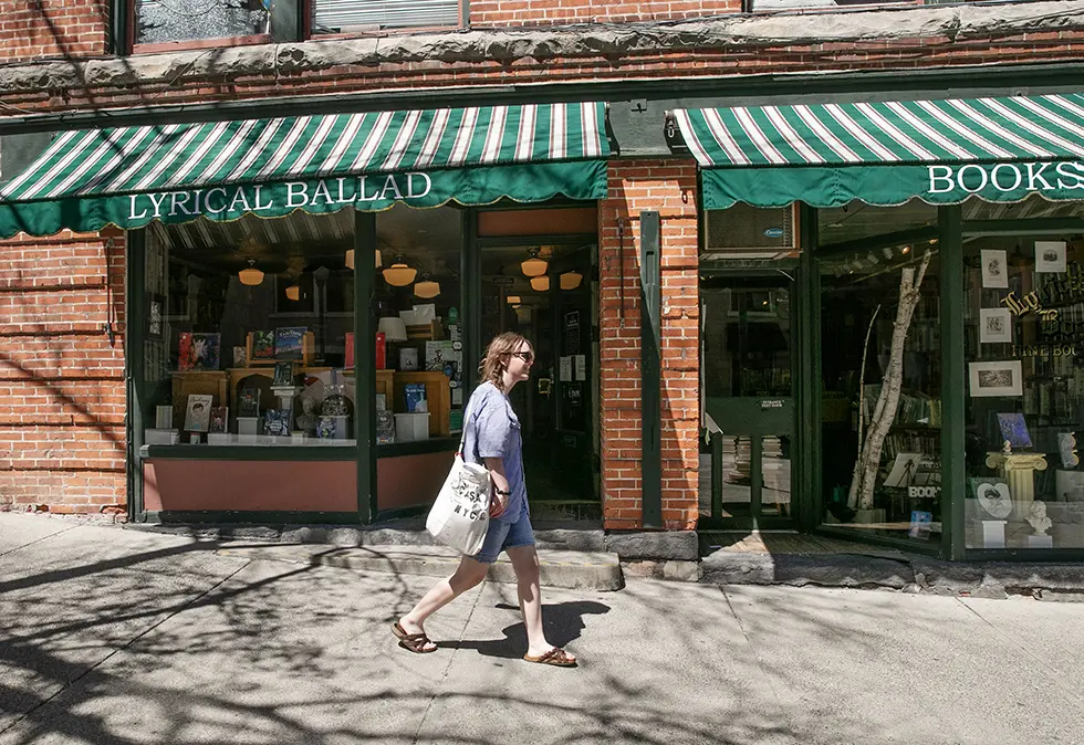 A student walks past Lyrical Ballad, a locally beloved independent bookstore in downtown Saratoga Springs, with a green-striped awning and inviting storefront display.