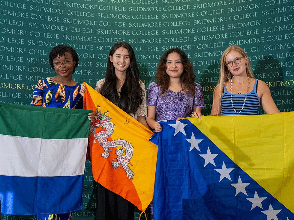 Four international students stand in front of a Skidmore backdrop holding their home flags.