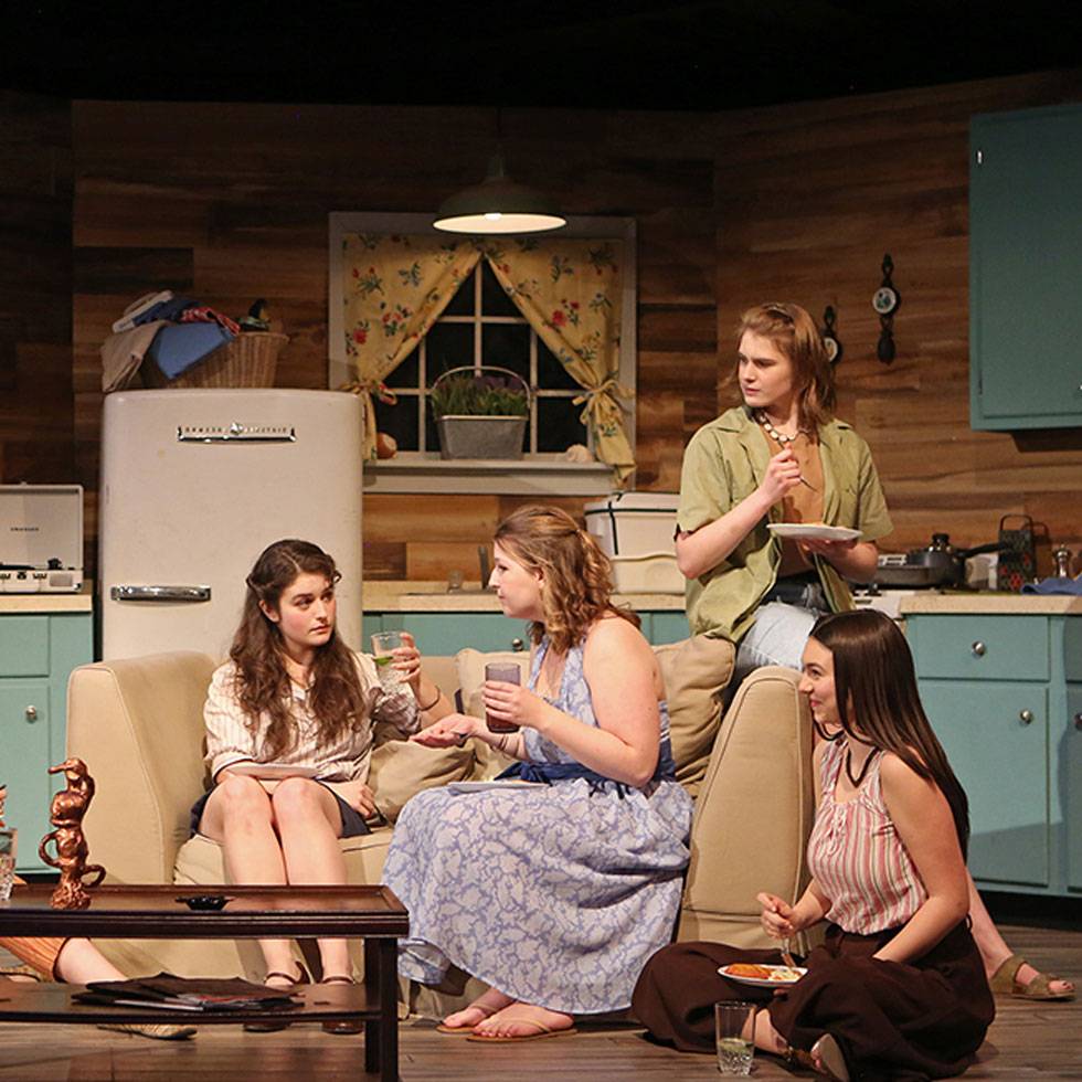 Four young women sit on a tan couch with a set designed kitchen in the background. All are focused on having a conversation with one individual in particular.