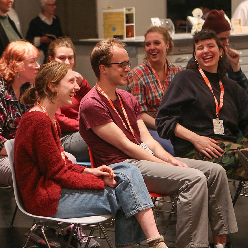A group of students sit in chairs smiling and laughing while facing forward.