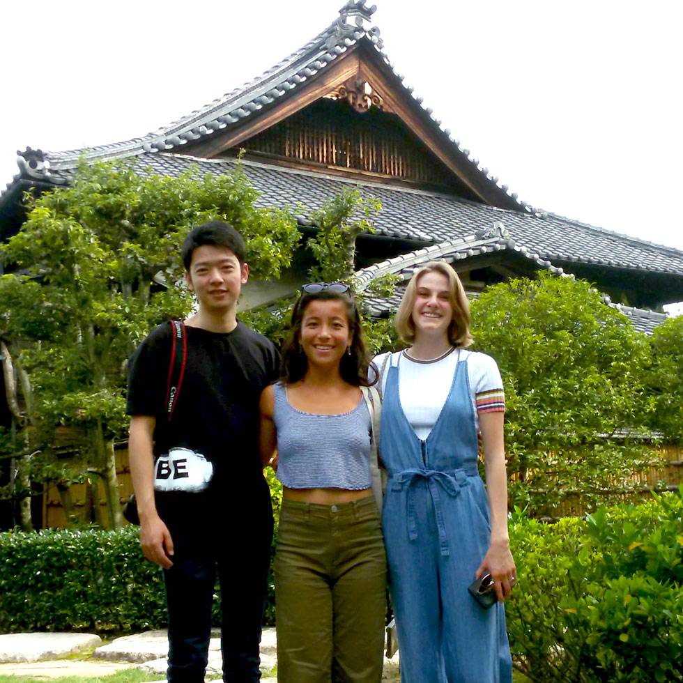 Three people stand close together, smiling at the camera in front of a traditional Japanese-style building with a tiled, curved roof. They are surrounded by lush green trees and shrubs, and one person holds a phone while another has a camera strap over their shoulder.